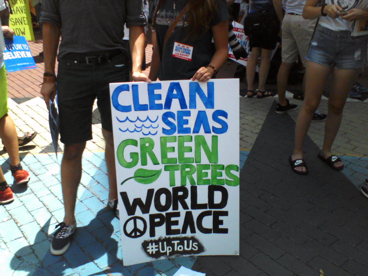 Handmade sign: "Clean Seas" in blue with blue waves, "Green Trees" in green with a green leaf, "World Peace" with a peace symbol in black and hashtag "#UpToUs".